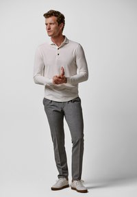 Weißes Langarm-Poloshirt mit drei Knöpfen, kombiniert mit grauen maßgeschneiderten Hosen und weißen Sneakers mit braunen Akzenten. Glatte Textur.