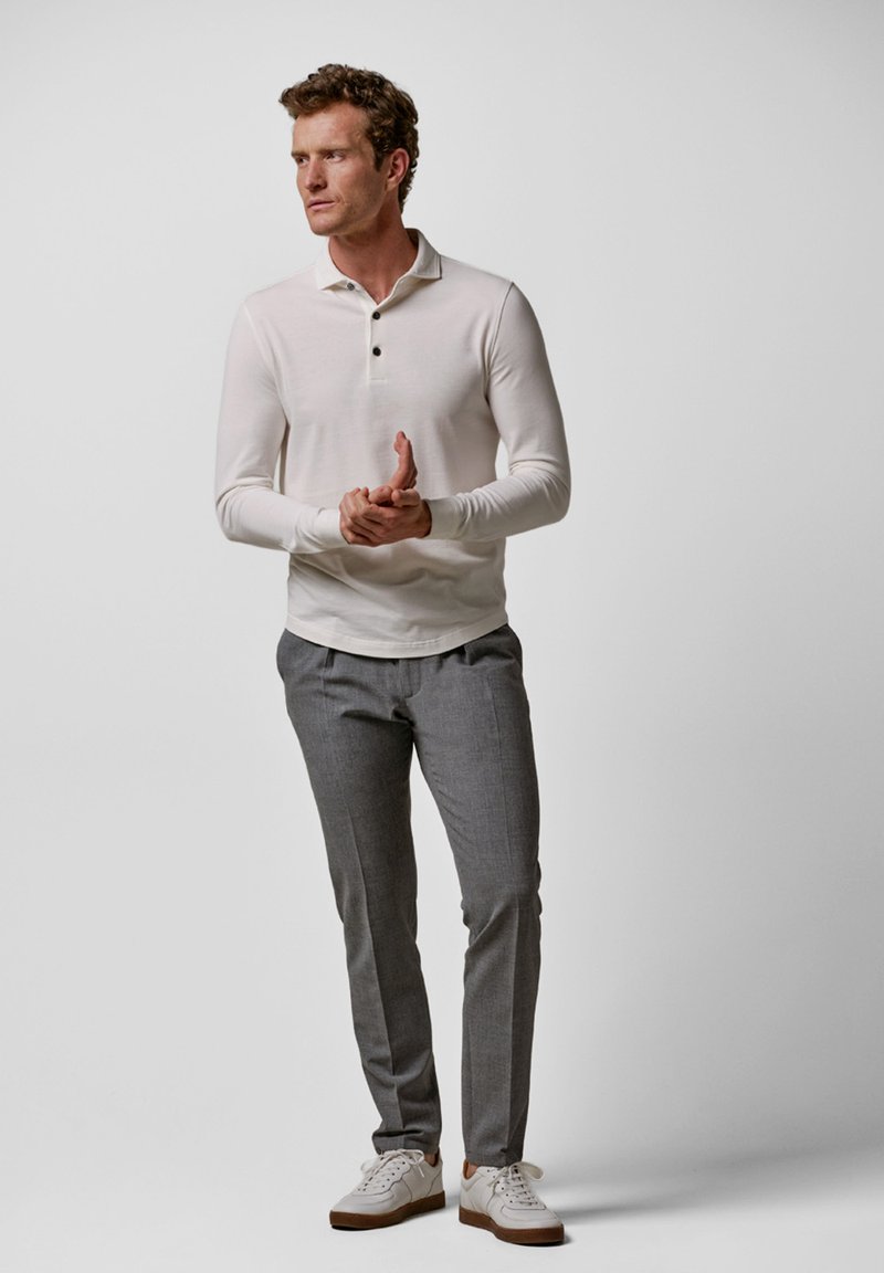 Weißes Langarm-Poloshirt mit drei Knöpfen, kombiniert mit grauen maßgeschneiderten Hosen und weißen Sneakers mit braunen Akzenten. Glatte Textur.