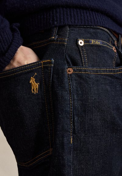 Χέρι στην πίσω τσέπη σκούρου μπλε τζιν με κίτρινες ραφές, με κεντημένο το λογότυπο Polo Ralph Lauren στην τσέπη.