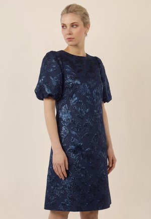 Woman wearing a navy blue, knee-length dress with puffed short sleeves and a shiny textured floral pattern, standing against a plain beige background.
