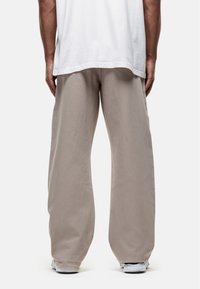 Beige wijde broek gemaakt van zachte katoen, met een relaxte pasvorm, gestikte details en een elastische tailleband. Witte sneakers zichtbaar onderaan.