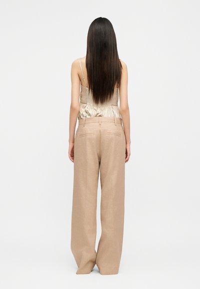 Γυναίκα με μακριά σκούρα μαλλιά που φοράει σατέν μπεζ τοπ με τιράντες και χαλαρά, καφέ παντελόνια με φαρδιά πόδια, γυρισμένη προς τα πίσω σε απλό φόντο.