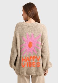 Pull en tricot beige surdimensionné avec un grand motif de fleur rose et orange et le texte "GOOD VIBES" au dos. Col rond, tissu texturé.