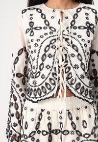 White embroidered blouse featuring black floral patterns and lace-up front. Long sleeves with scalloped edges; lightweight, breathable fabric.