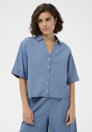 Kobieta z krótkimi kręconymi włosami w luźnej, jasnoniebieskiej koszuli z denimu zapinanej na guziki oraz pasujących szerokich spodniach, stojąca na białym tle.