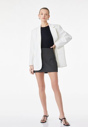 Donna con blazer bianco, top nero, gonna a pois bianco e nero e sandali neri con cinturini, in piedi con una mano sul fianco.