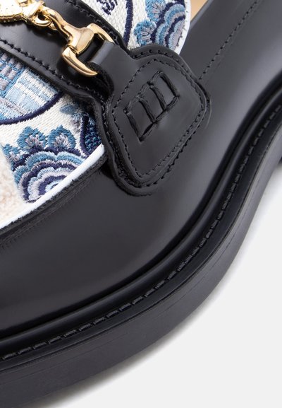Μαύρο δερμάτινο παπούτσι με χρυσά μεταλλικά στοιχεία και διακοσμητικό ύφασμα με μπλε λουλουδάτη κεντητική. Ομαλή υφή και στρογγυλεμένο σχήμα.