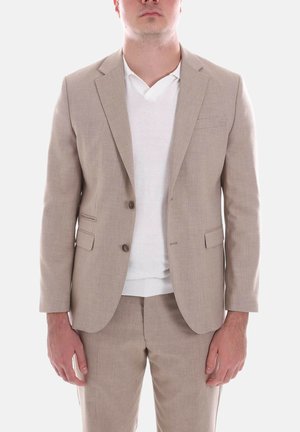 Uomo con giacca e pantaloni beige sopra una maglia bianca a scollo a V, in piedi con le braccia rilassate ai lati contro uno sfondo semplice.