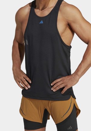 Man draagt een zwart mouwloos sporthemd en tweekleurige bruine en zwarte hardloopshorts, met handen op de heupen.