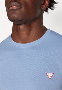 Lyseblå bomuld t-shirt med rund halsudskæring og et lille rødt triangulært "GUESS" logo på den øverste venstre bryst.