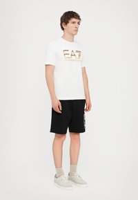 Hvit bomull t-skjorte med en metallisk gull "EA7" logo, sammen med svarte shorts med et sidemønster. Modellen har på seg lyse joggesko og sokker.