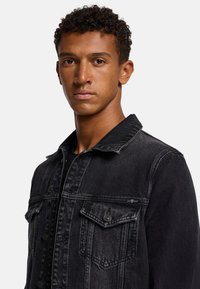 Svart denimjacka med knappstängning, två bröstfickor och krage. Har en sliten textur och subtil blekning.