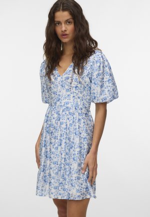 Femme aux longs cheveux ondulés portant une robe blanche mi-longue avec un imprimé floral bleu et des manches courtes bouffantes.