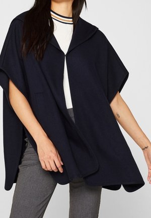 Vrouw in een marineblauwe cape-stijl jas over een wit gestreepte top en grijze broek, met zichtbare kleine tatoeages op haar armen.