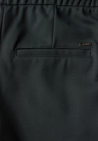 Tissu vert foncé avec une texture lisse, comportant une poche plate et une petite étiquette métallique cousue dans la couture latérale. Ceinture élastique visible.