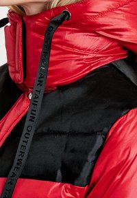 Czerwona kurtka puchowa z wysokim kołnierzem, kontrastującą czarną wstawką i teksturowanym materiałem. Posiada sznurek do regulacji z detalem marki.