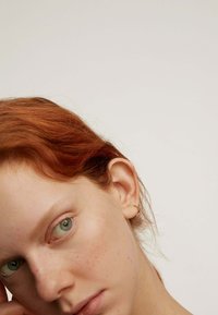 Primer plano de un rostro pálido con cabello rojo, ojos verdes y pecas ligeras. Un pequeño pendiente de aro de oro resalta en la oreja. Fondo blanco liso.