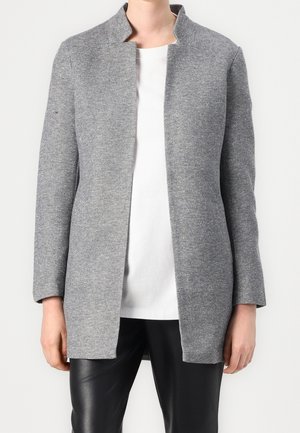 Persoon draagt een lange, grijze jas met open voorkant over een wit shirt en zwarte broek, staand tegen een effen achtergrond.