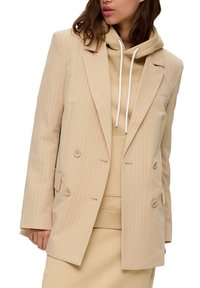 Žena v béžovém pruhovaném dvouřadém saku přes odpovídající mikinu s kapucí a sukní, s viditelným piercingem v septu.