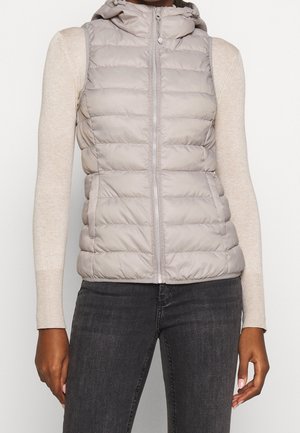 Kobieta w jasno szarej pikowanej kamizelce z kapturem, na beżowej bluzce z długim rękawem i ciemnoszarych dżinsach, stojąca na jednolitym tle.