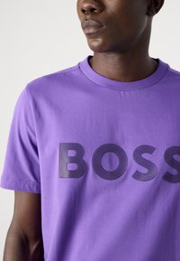 Fialové bavlněné tričko s kulatým výstřihem, s velkým černým texturovaným nápisem "BOSS" na hrudi. Hladká tkanina a krátké rukávy.
