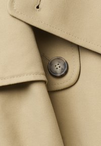 Bež trench coat kangas, millel on nööbidetail. Kanga pind on sile ja volditud, rõhutades nööbi ümmargust disaini ja õmblusi.