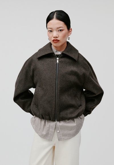 Blouson bomber en mélange de laine marron avec un grand col, fermeture éclair sur le devant et poches latérales, porté sur une chemise à rayures et un pantalon blanc.