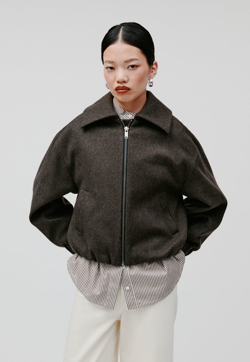 Giacca bomber in misto lana marrone con un grande colletto, chiusura zip anteriore e tasche laterali, indossata sopra una camicia a righe e pantaloni bianchi.