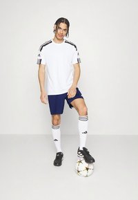 Hvit atletisk T-skjorte med svarte striper, marineblå shorts, hvite knestrømper med svarte detaljer og svarte fotballsko; spiller står på en fotball.