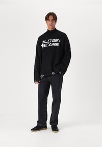 Μαύρο oversized πουλόβερ με λευκή εκτύπωση "LOVE SCARS", ριμπ λαιμό και τριμμένη στρώση στο τελείωμα. Συνδυάζεται με σκούρο τζιν και μαύρα αθλητικά παπούτσια.