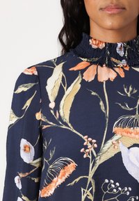 Floral navy blouse featuring a ruffled collar, long sleeves, and a mix of orange, white, and green botanical patterns on smooth fabric.