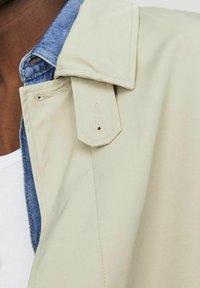 Beige jacka med en denimkrage, försedd med en knappslå, synliga sömmar och en mjuk textur. Lager på tyget för ökad djup.