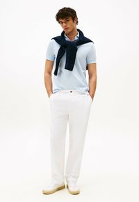 Polo shirt azzurro chiaro, maglione blu navy avvolto intorno al collo, pantaloni bianchi e sneakers bianche con suola beige.