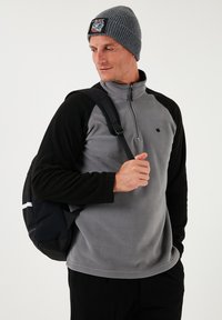Grå fleecepullover med sorte ærmer, kvart-zip krave, lille logo på brystet, parret med en grå strikket beanie og en sort taske.
