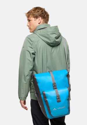 Man draagt een saliegroene capuchonjas en draagt een blauwgrijze Vaude schoudertas met gespflappen tegen een witte achtergrond.