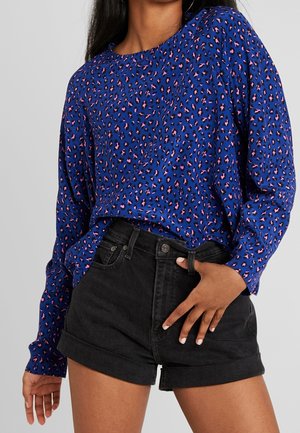 Femme portant un haut bleu à manches longues avec un imprimé léopard rose et noir et un short en jean noir taille haute avec des revers.
