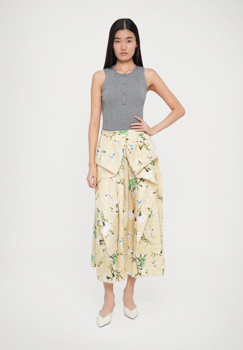 Grauer, taillierter Strickoberteil mit Knöpfen, kombiniert mit einem gelben, floral gemusterten Midikleid mit schichtigem Design und weicher Textur. Weiße spitz zulaufende Schuhe.