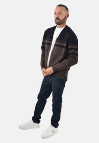 Cardigan blu scuro e marrone con righe orizzontali, indossato sopra una camicia bianca e abbinato a jeans scuri e sneaker bianche.