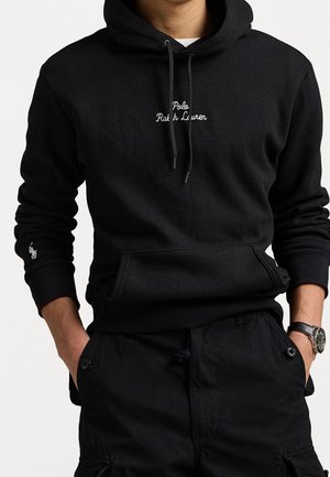 Osoba nosząca czarną bluzę z kapturem Polo Ralph Lauren z napisem logo na piersi i rękawie, ręce w kieszeniach oraz zegarek na lewym nadgarstku.