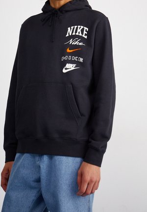 Osoba ubrana w czarną bluzę z kapturem Nike z wieloma białymi i pomarańczowymi logo Nike na piersi, w połączeniu z jasnoniebieskimi dżinsami.