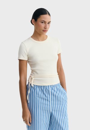 Femme portant un haut écru à manches courtes ajusté avec un nœud sur le côté et un pantalon à rayures verticales bleu et blanc, debout devant un fond uni.