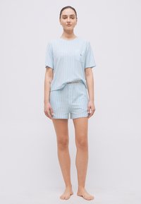 Γυναίκα που στέκεται ξυπόλητη, φοράει ανοιχτό μπλε και λευκό ριγέ μπλουζάκι με κοντά μανίκια και ταιριαστό σορτς με ορατό λογότυπο στη μέση.
