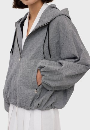 Person trägt einen grauen, strukturierten Kapuzenpullover mit Reißverschluss, schwarzen Kordeln und elastischem Saum, darunter ein weißes Hemd und einen weißen Rock.