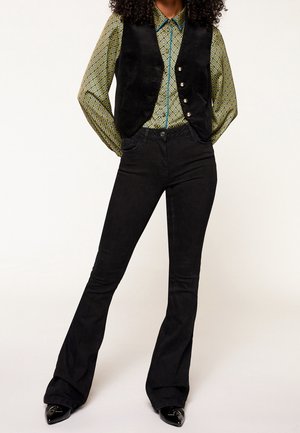 Vrouw draagt zwarte flarebroek, zwart aangeknoopt vest en een patroon lange mouwen overhemd, staand met handen achter de rug.