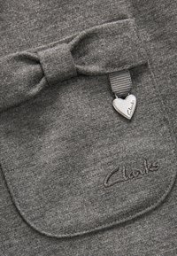 Grau genähtes Material mit glatter Textur zeigt eine Schleife als Akzent auf einer Tasche. Ein silbernes Herz-Anhänger mit "Clarks" und gesticktem Logo sind sichtbar.