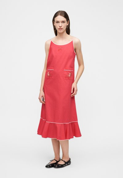 Γυναίκα που φοράει ροζ midi αμάνικο φόρεμα με κεντήματα λουλουδιών στις τσέπες και βολάν στο κάτω μέρος, συνδυασμένο με μαύρα Mary Jane παπούτσια με καρφιά.