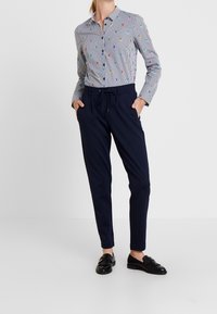 Kvinna iförd marinblå tapered byxor, svarta loafers och en långärmad randig skjorta med små färgglada fågelmotiv, med händerna i fickorna.