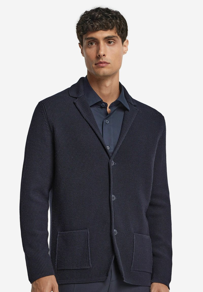 Marineblå strikket cardigan med et struktureret mønster, klippet krave, tre knapper og to frontlommer. Fremstillet af et varmt, strækbart materiale.