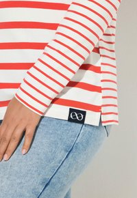 Rot-weiß gestreiftes Langarmshirt aus weicher Baumwolle, mit einem Seitenschlitz und einem navyblauen Etikett mit einem Symbol am Saum.