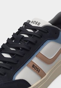 Gros plan sur une basket BOSS avec des lacets noirs, des accents bleu marine et brun clair, et le logo BOSS sur la languette et le côté.